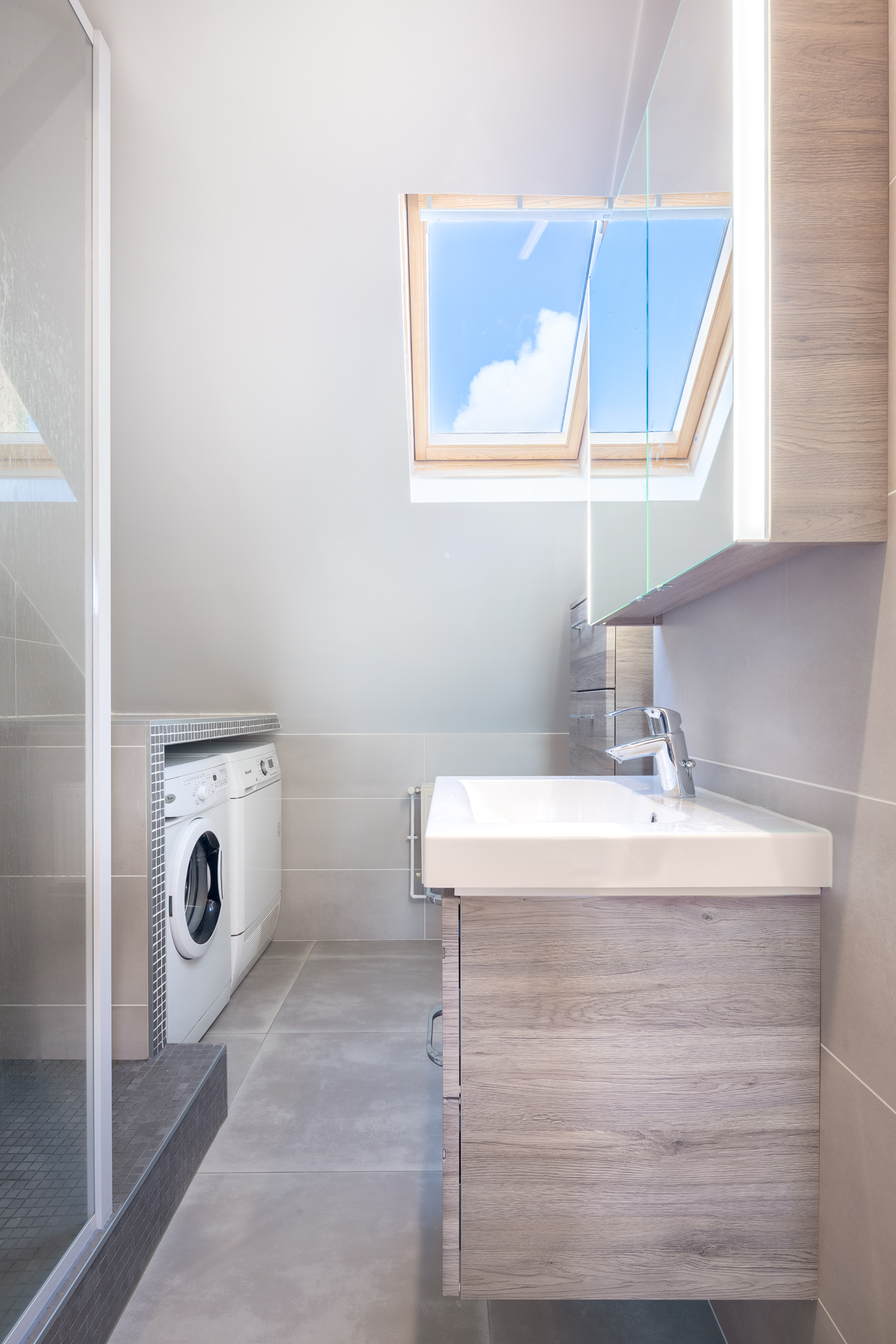 Vue de salle de bain toute en profondeur avec un lavabo, des placards, les machines pour le linge, une douche. Une fenêtre velux fait rentrer la lumière du jour avec un grand ciel bleu.